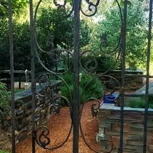 Close-up of a decorative wrought iron gate with swirling patterns, opening to a garden with gravel path and stone walls.